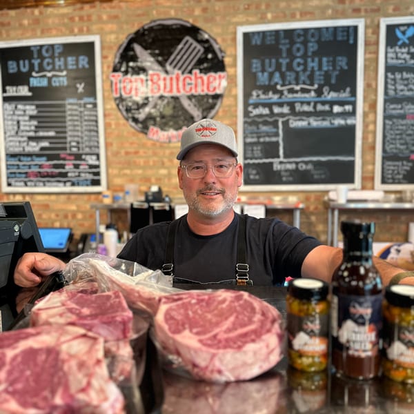 A smiling butcher stands behind a counter with fresh cuts of meat and Top Butcher Market sauces.