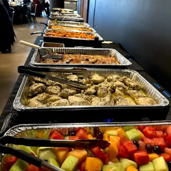 A buffet line with trays of chicken, pasta, and fresh fruit salad at Top Butcher Market.