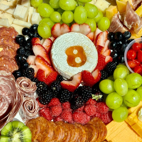 A vibrant charcuterie board with cheeses, grapes, berries, salami, prosciutto, and a round of brie with honey.