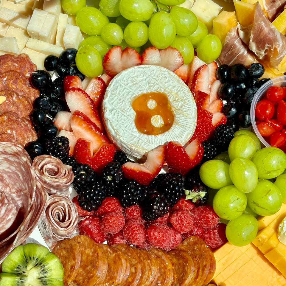 A vibrant charcuterie board with cheeses, grapes, berries, salami, prosciutto, and a round of brie with honey.