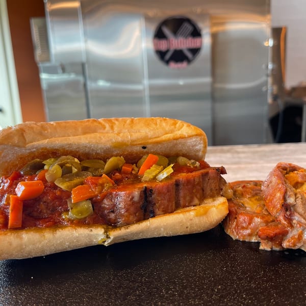 A Top Butcher Market sausage sandwich with peppers and sauce, next to a piece of sausage.