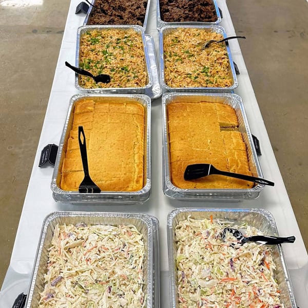 Catering trays of pulled pork, mac and cheese, cornbread, and coleslaw on a white tablecloth.